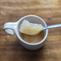 A spoonful of bone broth is shown resting on the edge of a white mug, with the broth dripping slightly onto the wooden surface below.